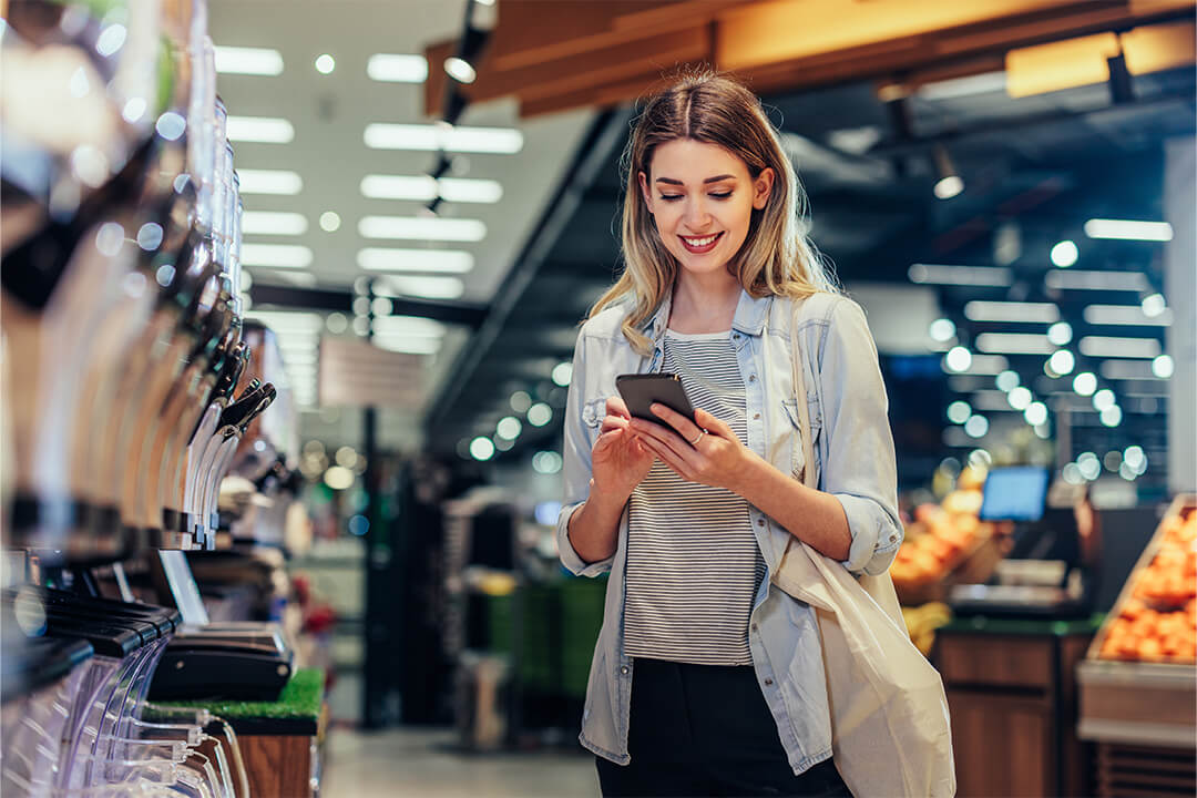 Lächelnde Frau mit Einkaufstasche in einem Supermarkt, die auf ihr Smartphone schaut. 
