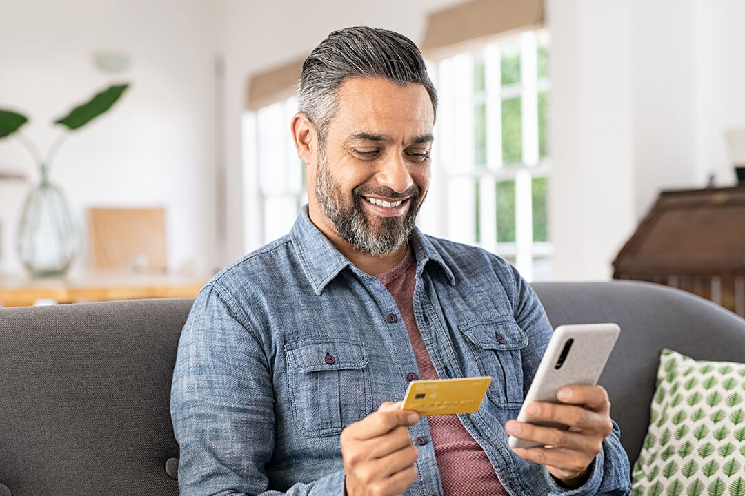 Ein Mann sitzt auf einem Sofa und hält ein Smartphone und eine Kreditkarte in der Hand, um online einzukaufen.