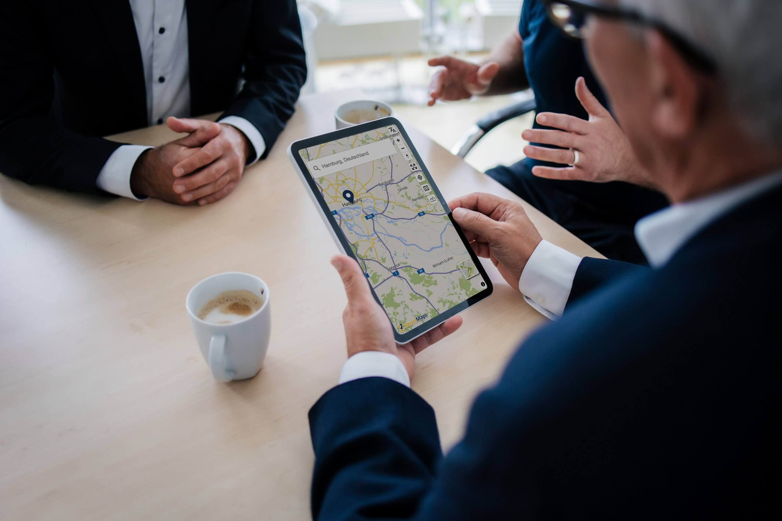 Mehrere Personen sitzen an einem Tisch, eine Person hält ein Tablet mit einer SmartMaps-Karte auf der ein geokodierten Standort zu sehen ist.