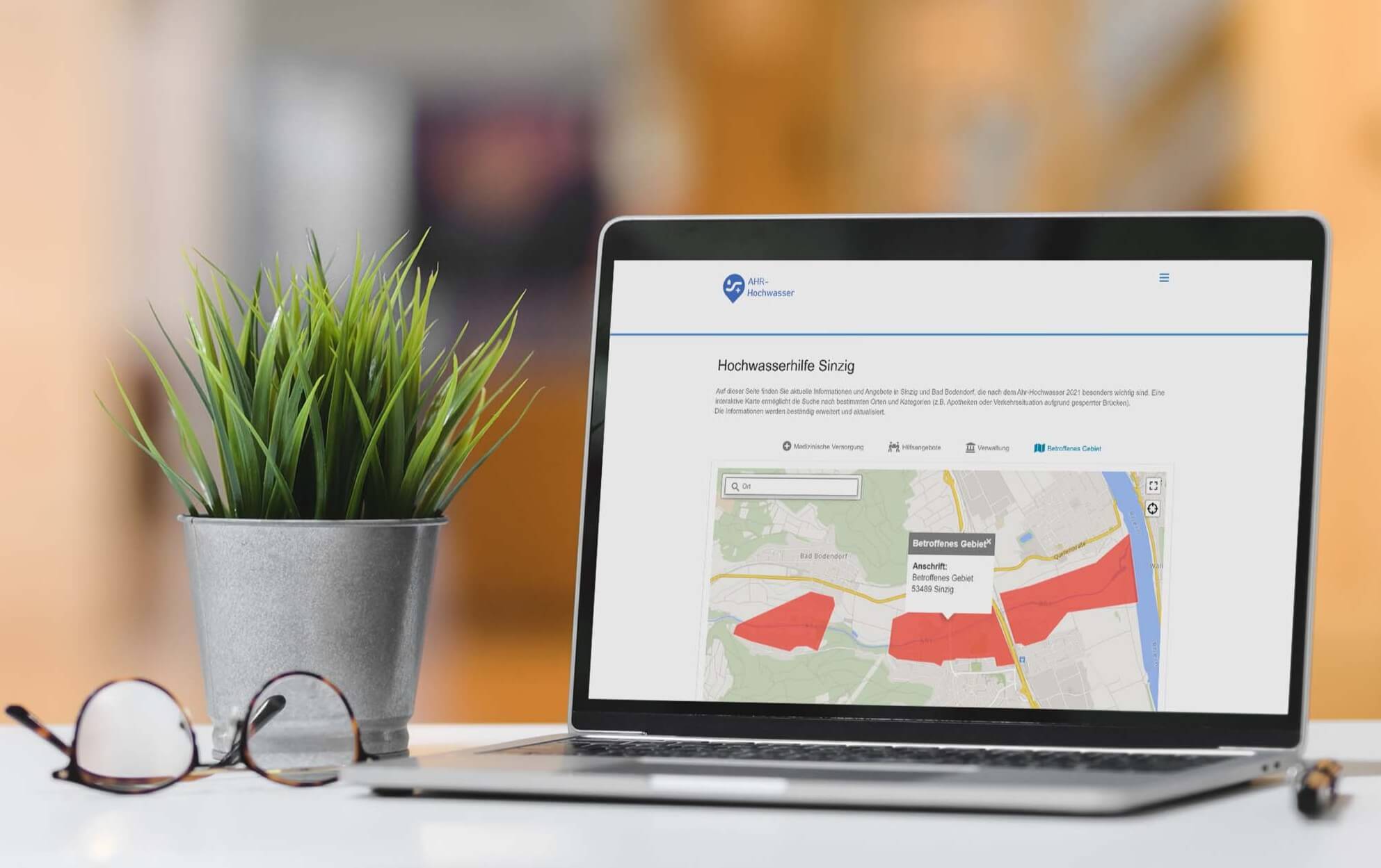 Auf einem Schreibtisch steht ein Laptop mit der SmartMaps-Anwendung der Hochwasserhilfe Sinzig. Auf dem Bildschirm ist ein Kartenausschnitt von Sinzig mit markierten Überflutungsgebieten und einem Anzeigefenster mit Informationen zum ausgewählten Gebiet zu sehen.