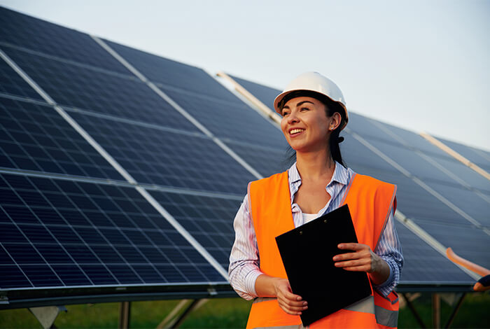 Eine Frau in orangefarbener Warnweste und Schutzhelm mit einem Clipboard in den Händen steht vor einer Reihe von Solarpanelen.