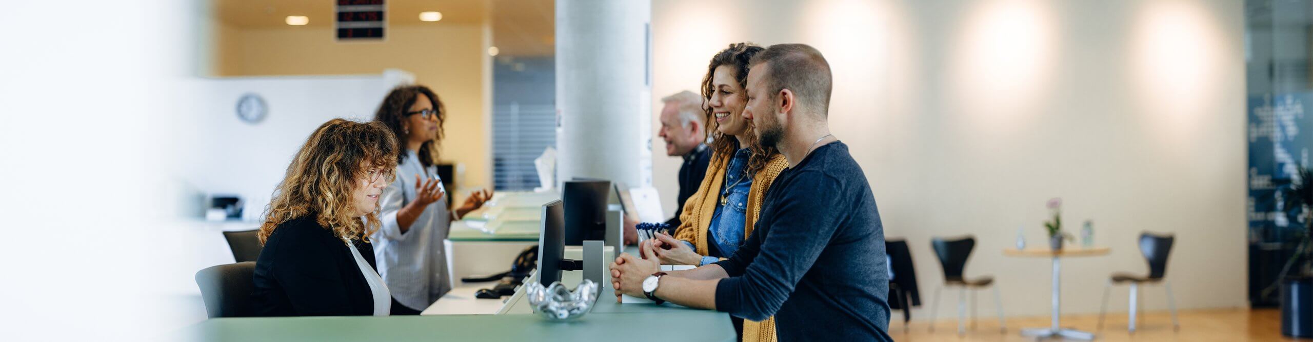 An einem Empfangsschalter in einem Amt stehen drei Menschen. Die Besucher werden von zwei Mitarbeitenden, die an Computern arbeiten, beraten.